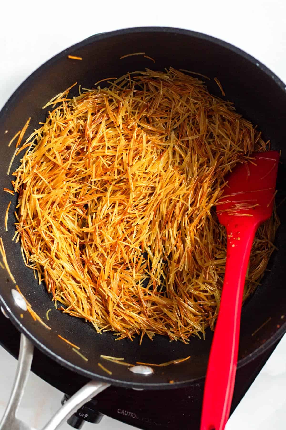 Toasted vermicelli noodles and spices in a pan with a red spatula.