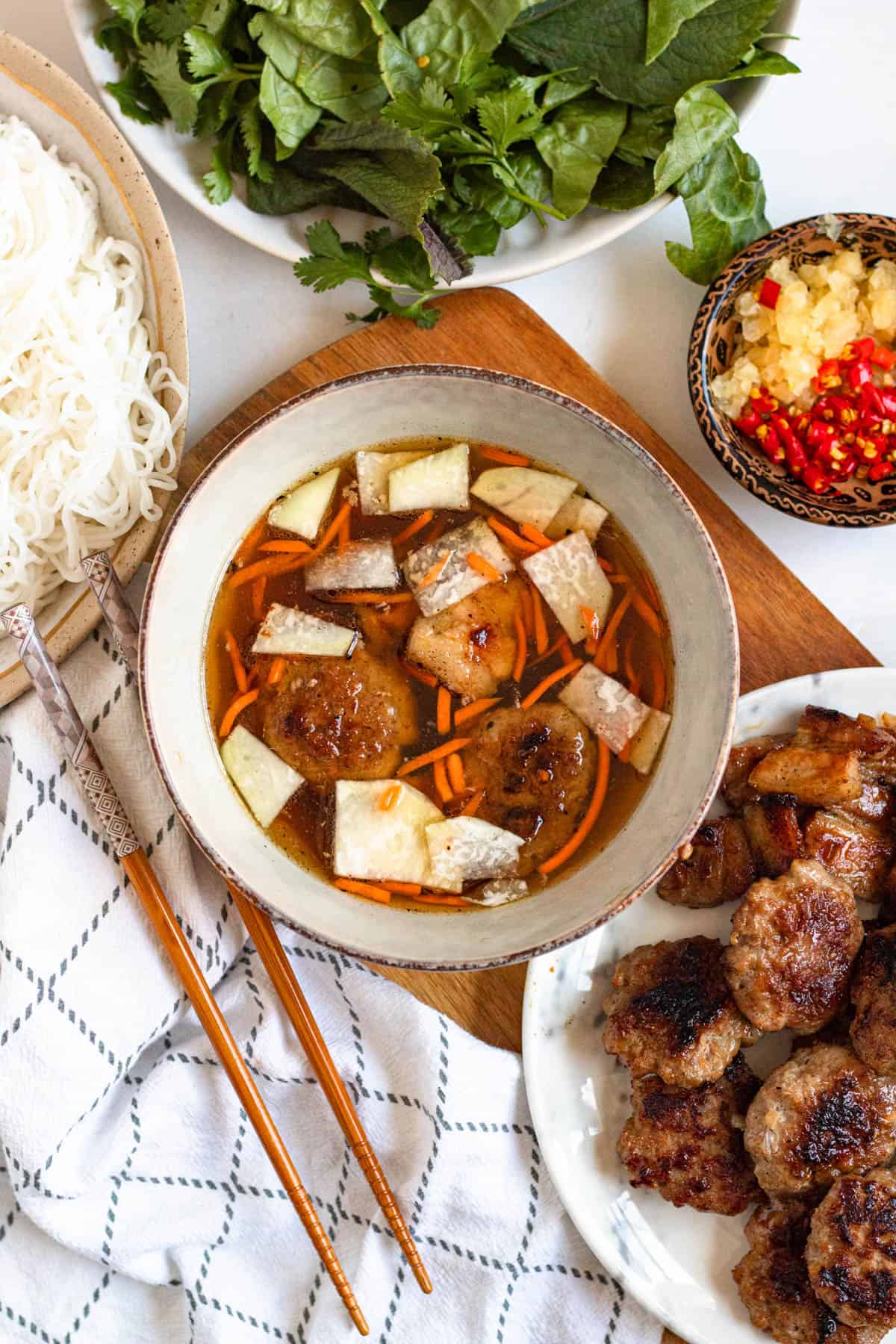 Broth with meatballs and vegetables in it, with vermicelli noodles, chopsticks, and chilies on the side.
