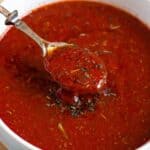 Rich homemade pizza sauce with Italian herbs scooped onto a spoon, ready to spread on pizza dough.