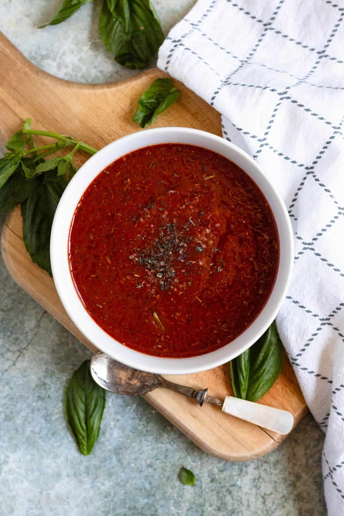 Homemade pizza sauce in a white bowl with herbs and a spoon on the side.