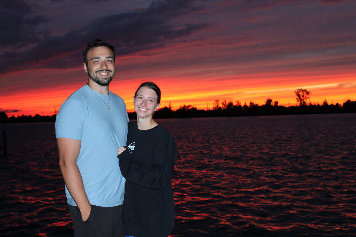 A couple smiling in front of an orange sunset. 