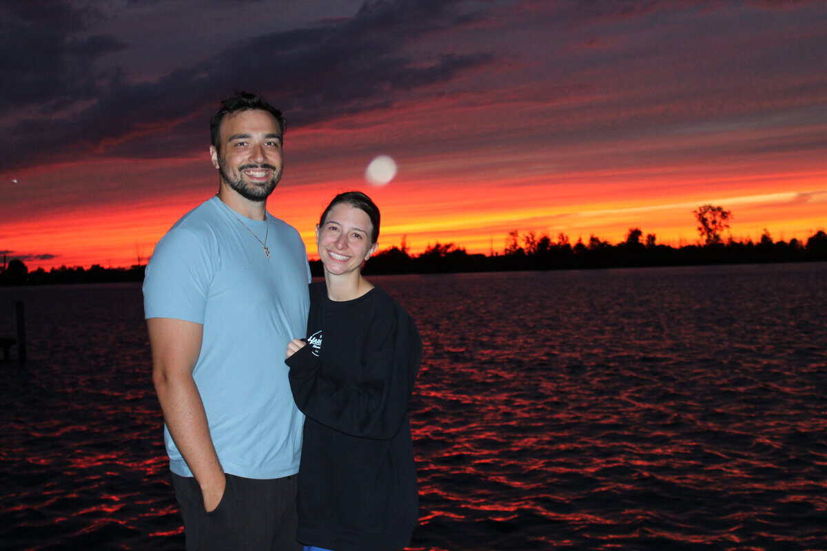 A couple smiling in front of an orange sunset with an orb of light next to them. 